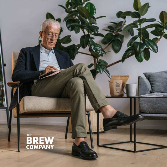 elderly man reading book about barista coffee techniques sitting in modern living room with coffee package and cup on table